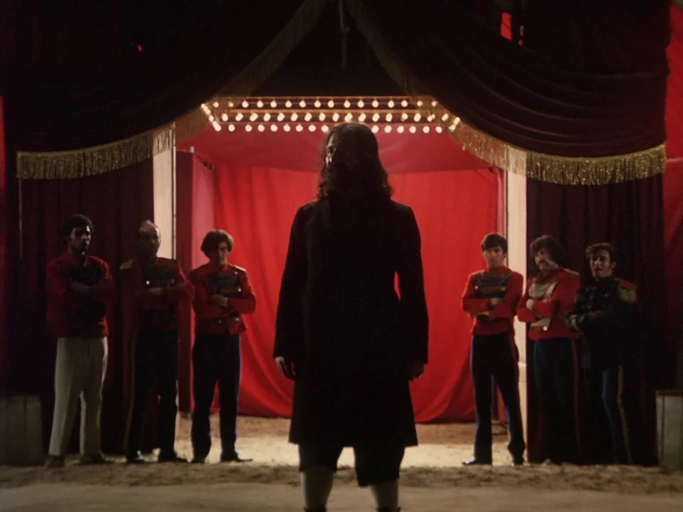 The Clowns (1970) movie still: fullbody — A dark arena features a large red curtain backdrop illuminated by a string of bulbs. A central bearded man…