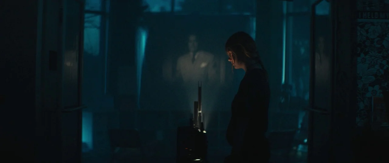 I Think We’re Alone Now (2018) movie still: medium — A dark, blue-toned room features a central projector casting an image onto a screen. A young…
