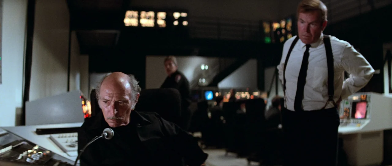 Escape From New York (1981) movie still: medium — A dimly lit industrial operations center contains multiple tiered computer consoles and control…