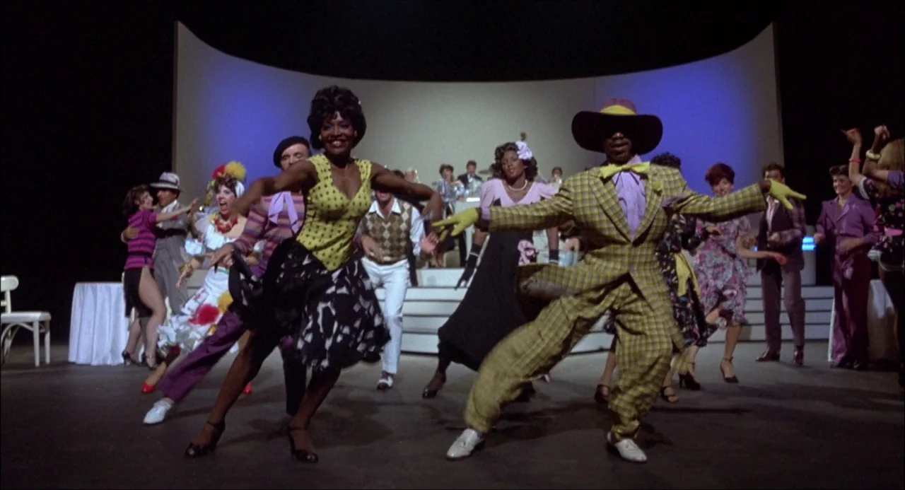 Xanadu (1980) movie still: fullbody — A spacious dance hall with a curved white wall and a tiered bandstand background. Dancers in period-accurate…