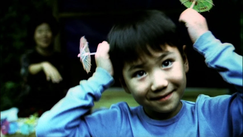 Save The Green Planet! (2003) movie still: closeup — A young boy plays in an outdoor space while a woman sits blurred behind. The boy holds two…