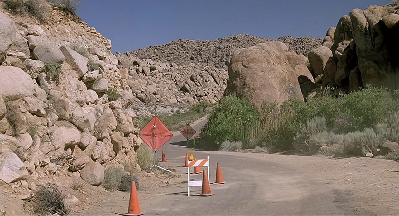 Tremors (1990) movie still: establishing — A winding paved road cuts through a canyon of large, sun-baked desert boulders. Orange traffic cones and…