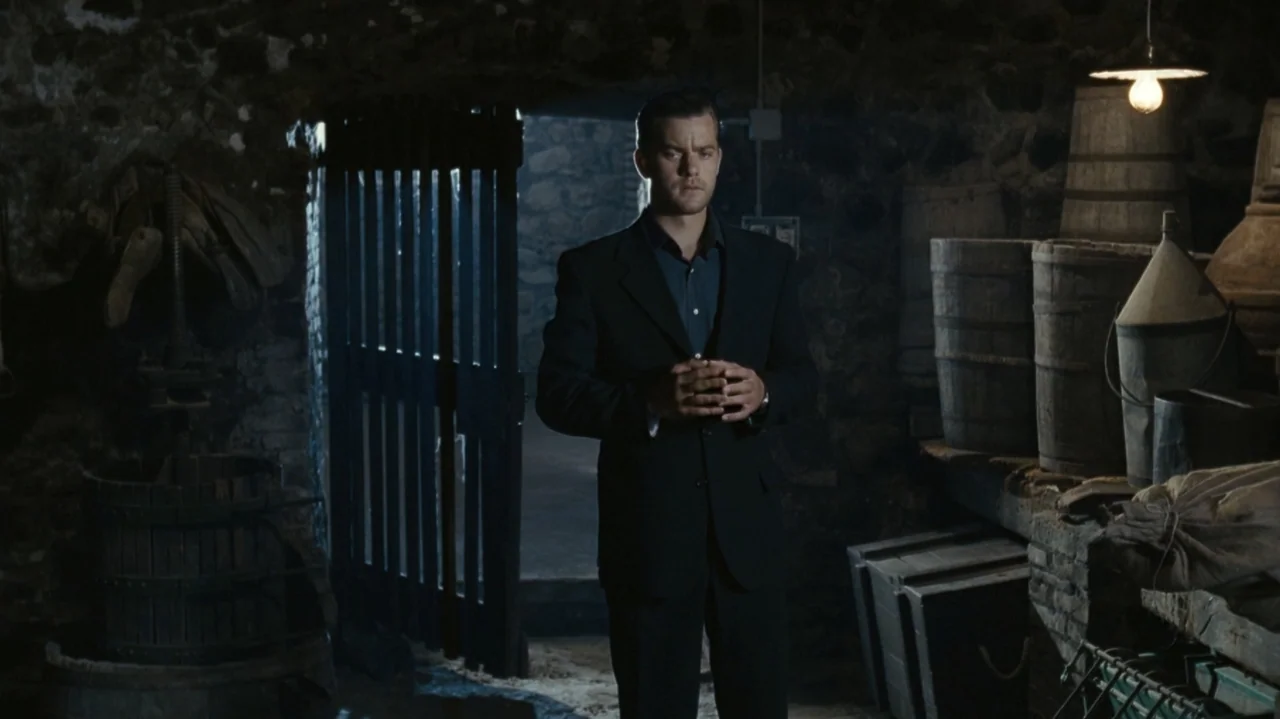 Shadows in the Sun (2005) movie still: medium — A stone-walled cellar features wooden barrels and a gate leading to a dark area. A man in a dark…
