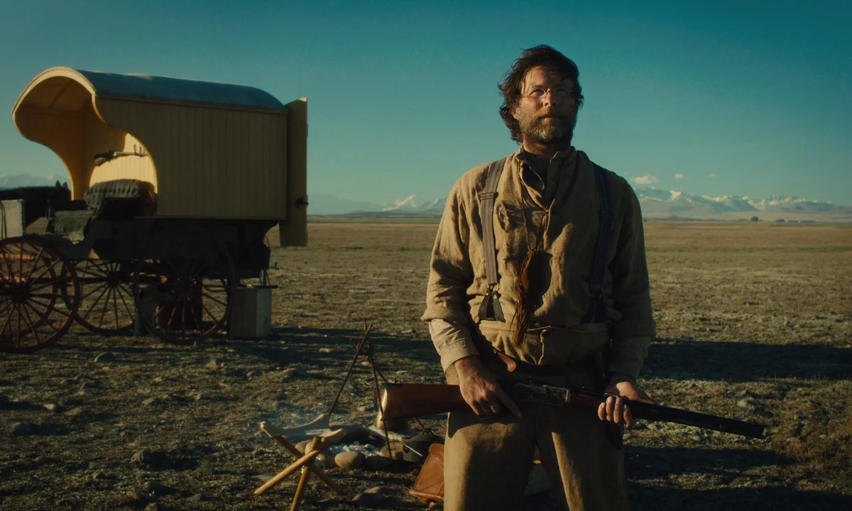 Slow West (2015) movie still: fullbody — A vast, dry prairie landscape stretches toward distant snow-capped mountains under a clear sky. A man…