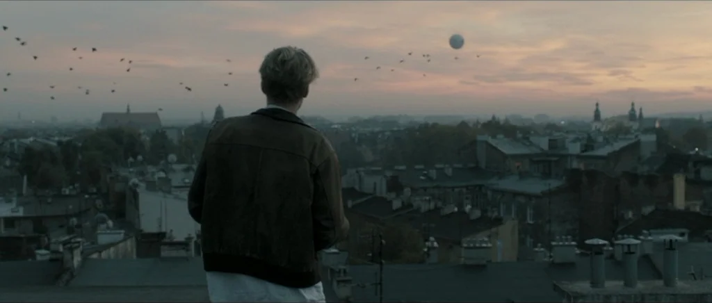 Lasting (Nieulotne) (2013) movie still: medium — A view across a dense urban rooftop landscape during a hazy dusk. A young man stands in the…
