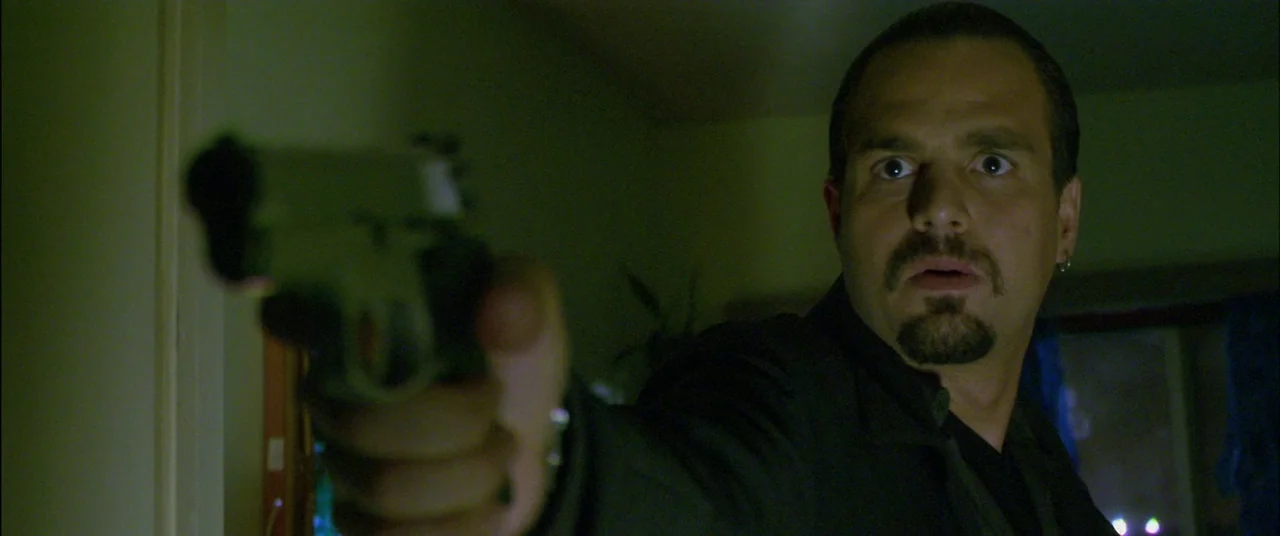 Collateral (2004) movie still: closeup — An dimly lit interior space with shadows obscuring the surroundings. A man with a goatee holds a handgun…