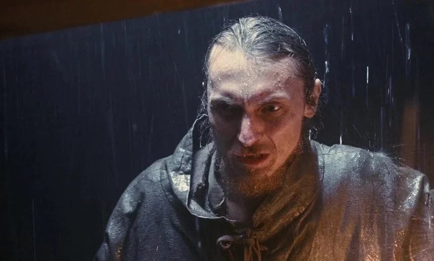 Nosferatu (2024) movie still: closeup — A man stands outdoors in heavy rain during a dark night. The individual is wet and stares forward with an…
