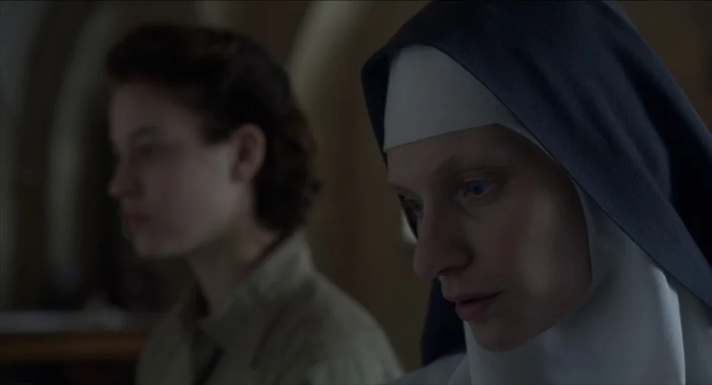The Innocents (2016) movie still: medium — A dimly lit interior space features a nun in foreground and woman behind. The nun wears a habit while the…