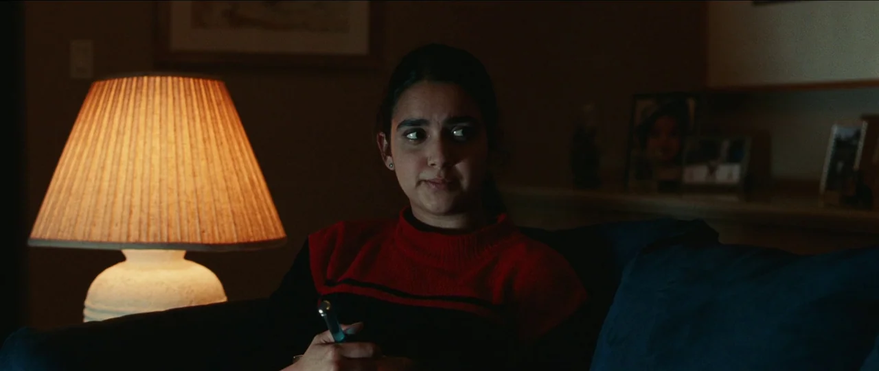 Bad Education (2019) movie still: medium — A dimly lit room with a table lamp providing the primary light source. A woman sits on a dark sofa while…