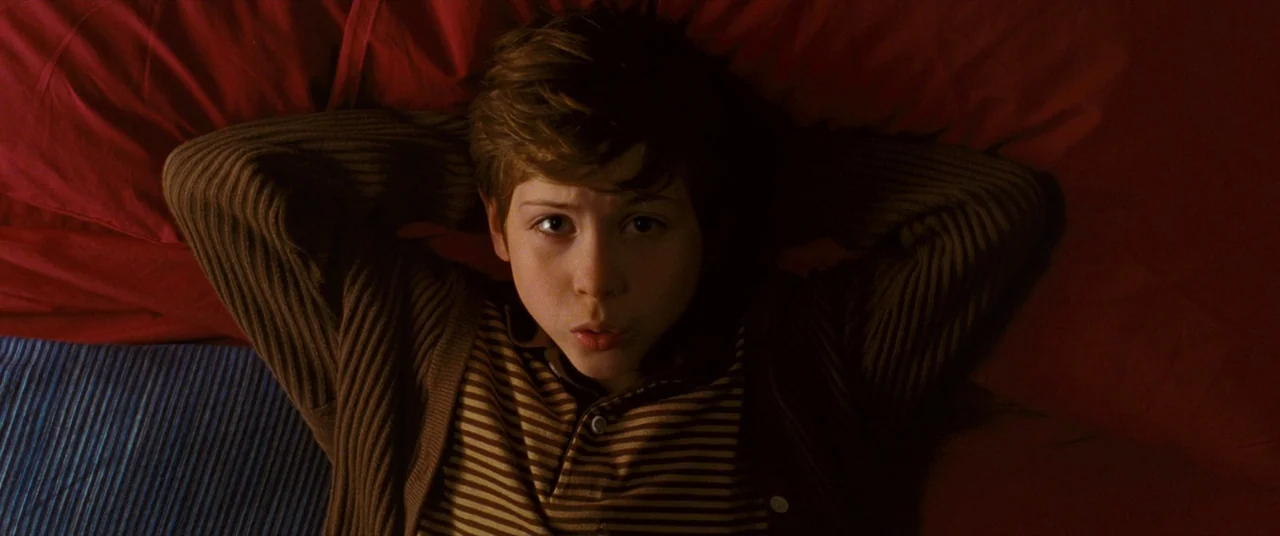 The Death and Life of John F. Donovan (2018) movie still: closeup — A boy lies on a bed with red sheets and a blue textured blanket. He rests with…
