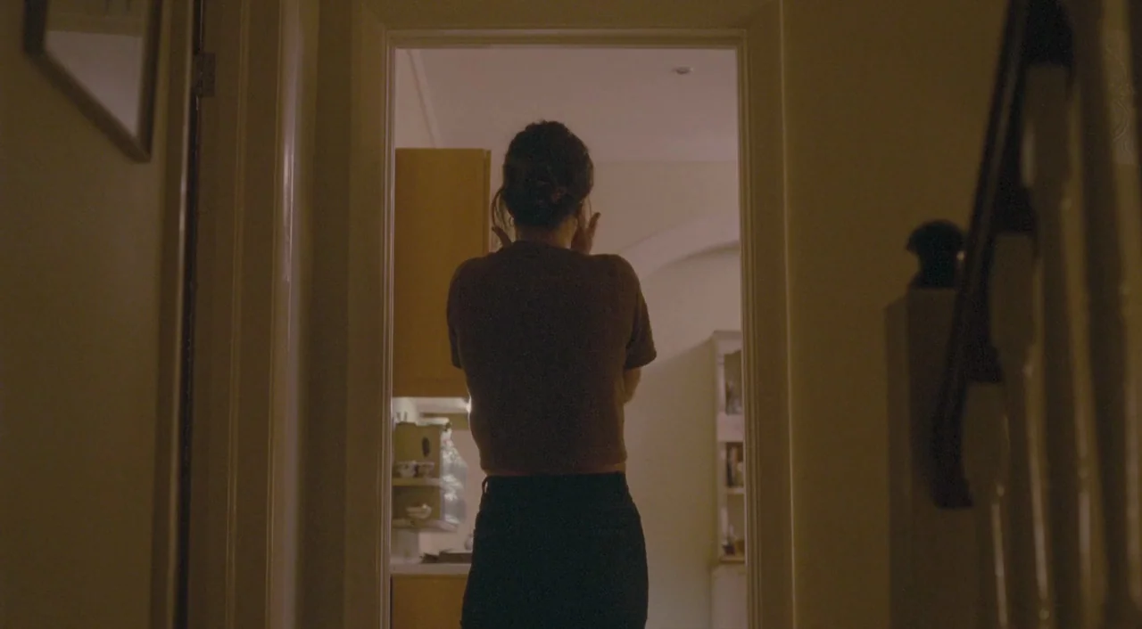Broken (2012) movie still: medium — A person stands in a doorway between two rooms in a home. The figure faces away from the camera with hands held…