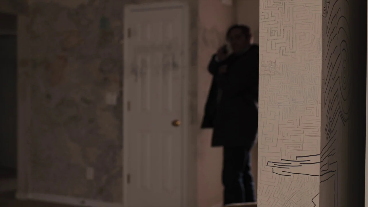 Prisoners (2013) movie still: medium — A dimly lit indoor space features a wall covered in intricate, hand-drawn marker designs. A man stands in the…