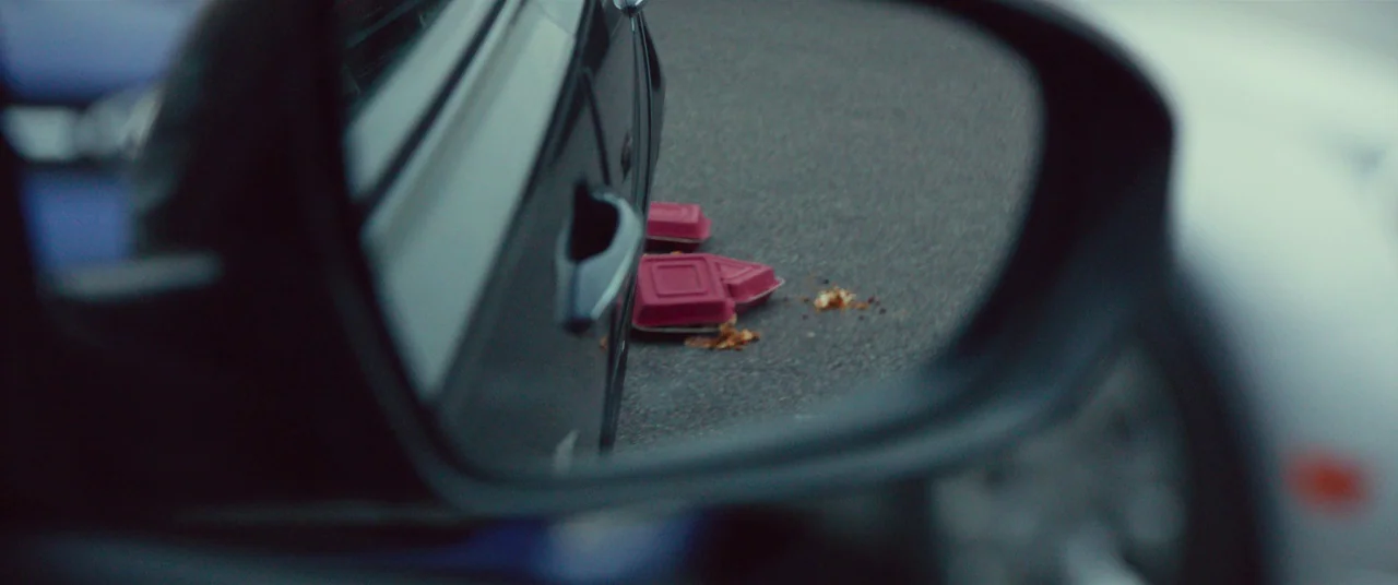 Split (2016) movie still: closeup — Two crushed red plastic food containers sit on gray asphalt. They rest beside the lower body of a dark-colored…