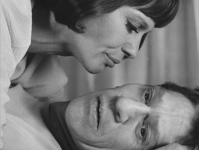 The Rite (1969) movie still: closeup — A close-up of two people lying in bed with white curtain fabric in background. The woman leans over the man…