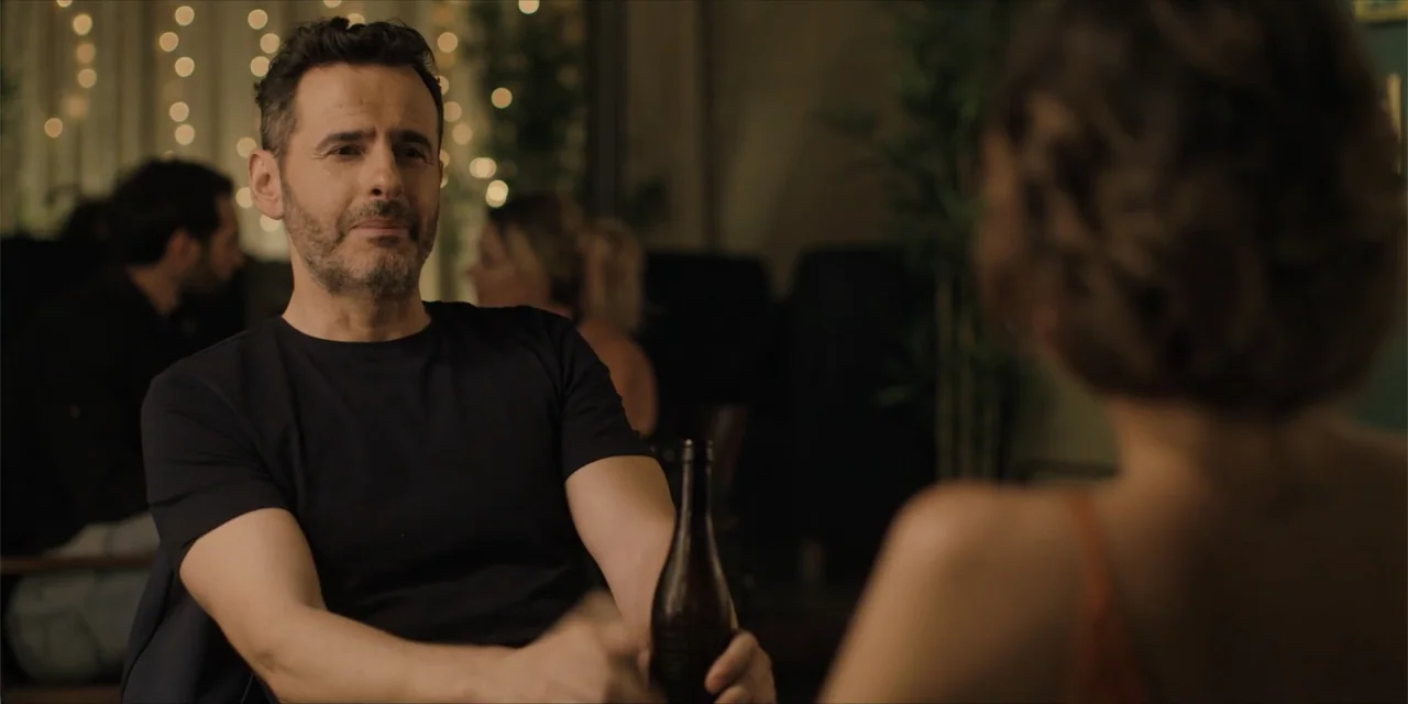 Instant Love (2020) movie still: medium — A dimly lit indoor space featuring string lights and blurred figures in the background. A man in a dark…