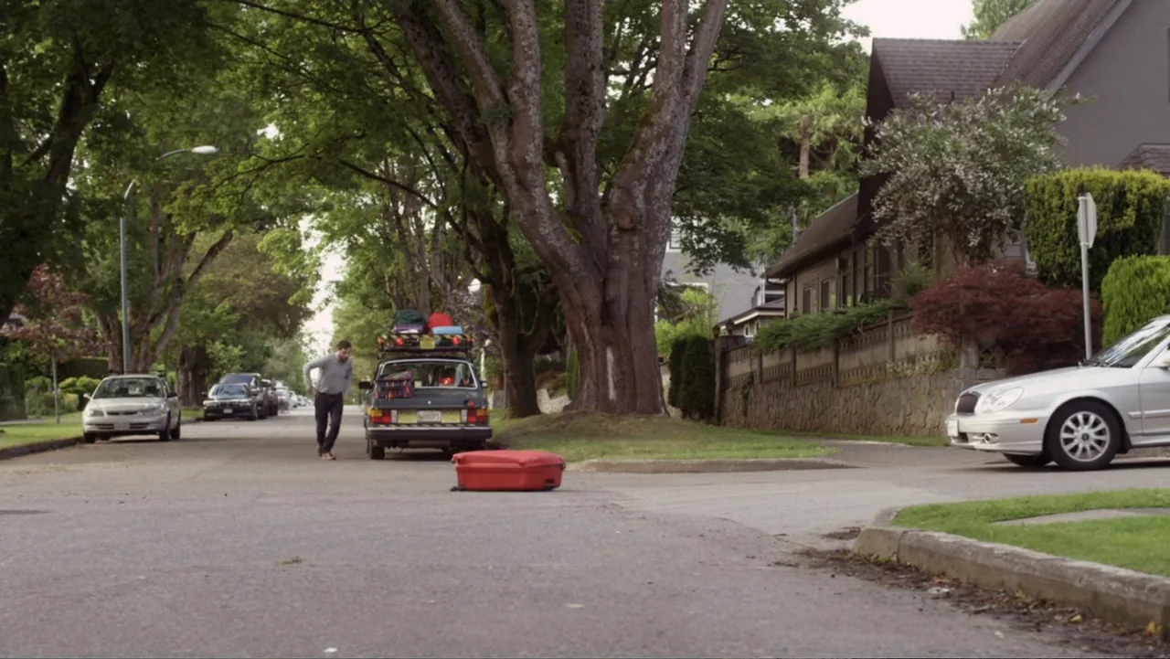 Jinxed (2013) movie still: wide — A suburban residential street features mature trees, parked cars, and houses along the sidewalks. A man runs…