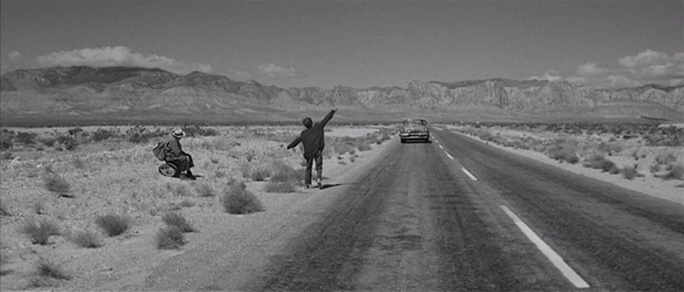 In Cold Blood (1967) movie still: wide — A vast, flat desert landscape spans the frame beneath rugged, arid mountains. Two figures wait by the side…
