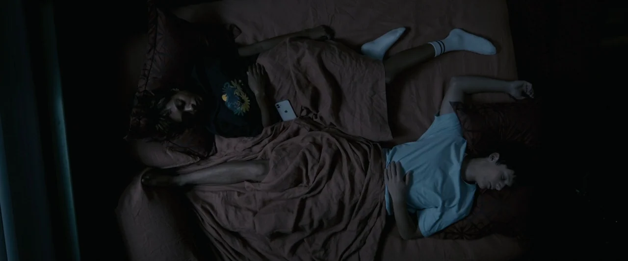 Talk to Me (2023) movie still: fullbody — Two individuals are lying asleep in a bed under a brown cover. One person is positioned on the left and…