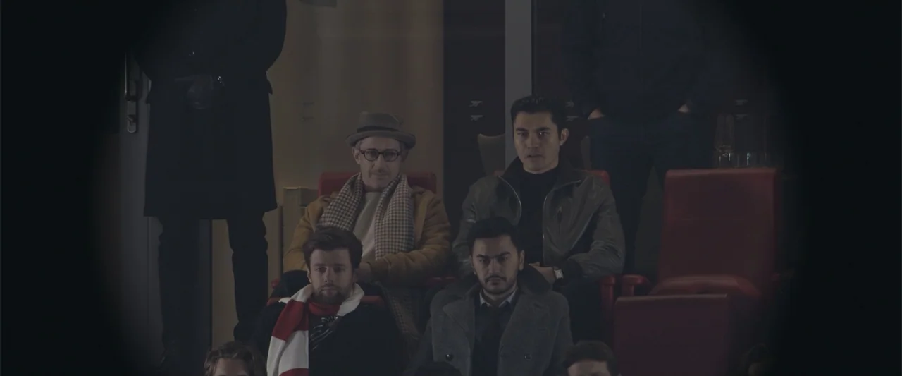 The Gentlemen (2020) movie still: medium — A dimly lit stadium suite features rows of red chairs and obscured figures behind. Four men sit in the…