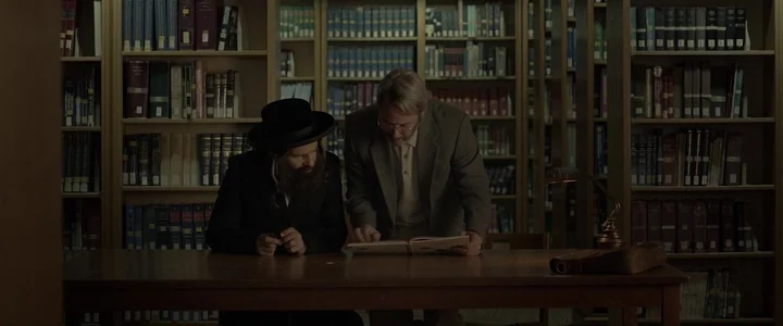 To Dust - Nightmares (2018) movie still: medium — A quiet library with rows of densely packed bookshelves lining the background. Two men stand at a…
