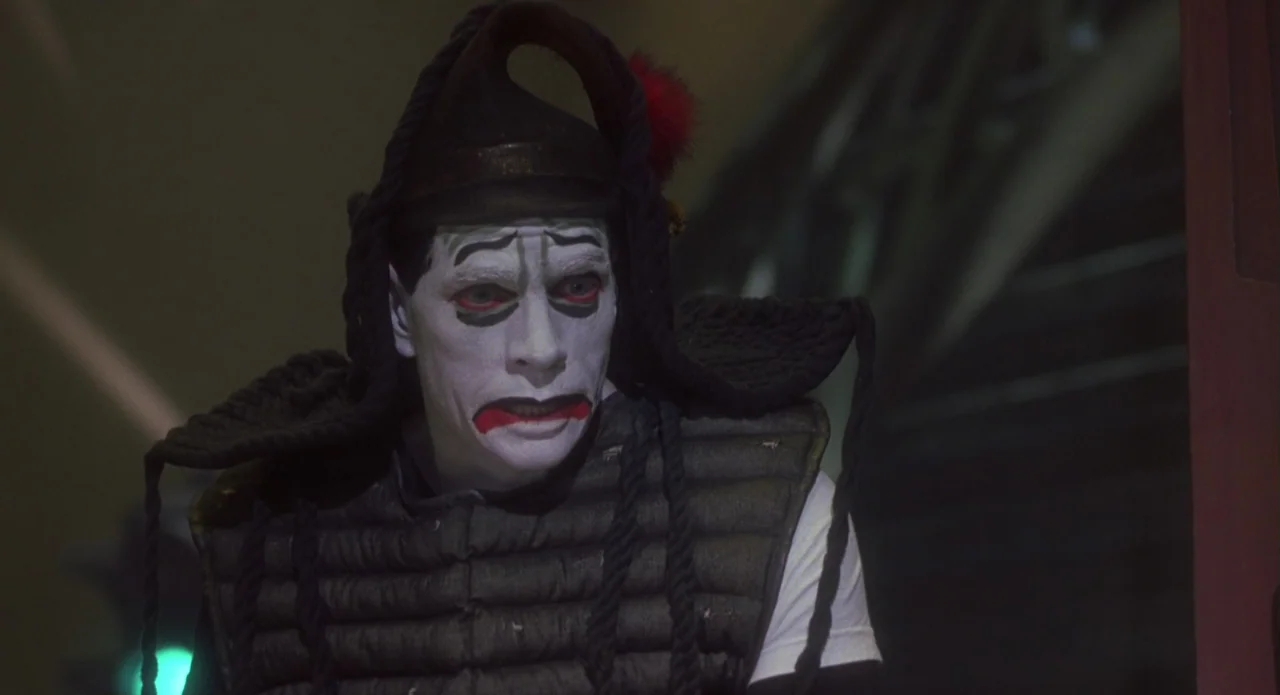 Monkeybone (2001) movie still: closeup — A dimly lit indoor space features a person dressed in theatrical circus attire. The individual wears heavy…