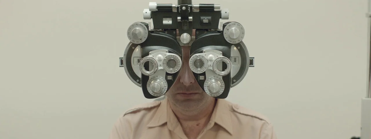 Entertainment (2015) movie still: closeup — A clinical examination room features stark off-white walls and neutral medical equipment. A man sits…