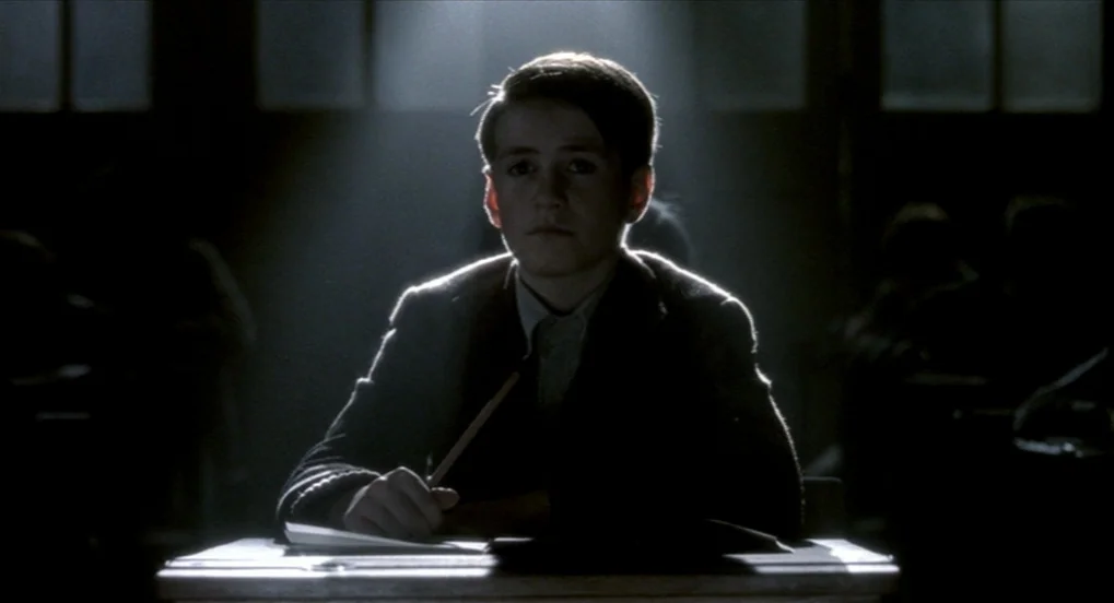 The Long Day Closes (1992) movie still: medium — A dimly lit classroom features rows of wooden desks arranged in a formal layout. A young boy in a…