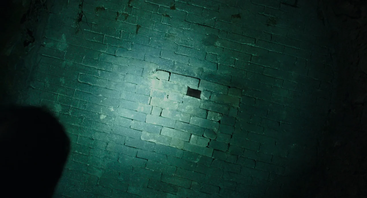The Mimic (2017) movie still: closeup — A dark, curved brick wall is partially lit by a concentrated, narrow light source. The beam highlights a…