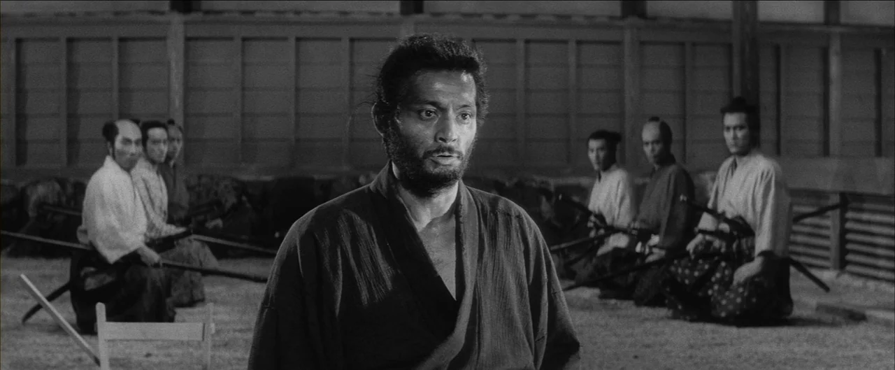 Harakiri (1962) movie still: medium — A spacious training hall with wooden paneling and a sand-covered floor. A man stands in the foreground as…
