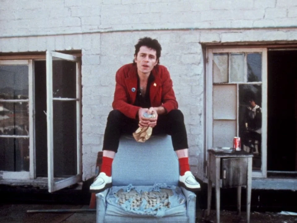 Decline of Western Civilization (1981) movie still: fullbody — A man sits on a worn, tattered armchair placed against a white brick wall. Two…