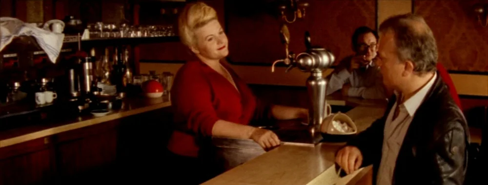 Carne (1991) movie still: medium — A dimly lit bar with wood paneling and a stainless steel draft beer tap. A woman behind the bar speaks to a man…