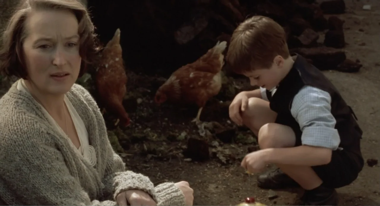 Dancing at Lughnasa (1998) movie still: medium — An outdoor farmyard features dirt ground with a woman and a child sitting nearby. The woman watches…