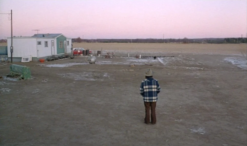 Stroszek (1977) movie still: wide — A flat, dusty rural lot features a small white mobile home and sparse clutter. A lone man stands in the…