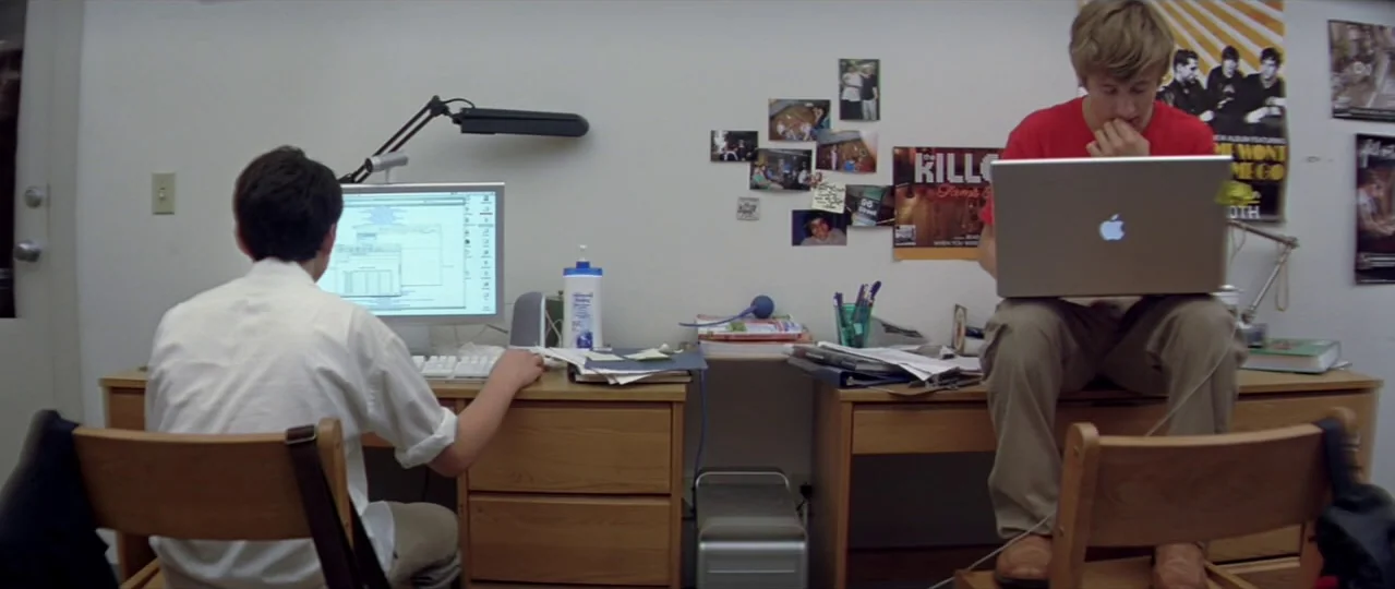 Afterschool (2008) movie still: wide — A college room with two wooden desks and white walls covered in small photos. Two young men work on their…