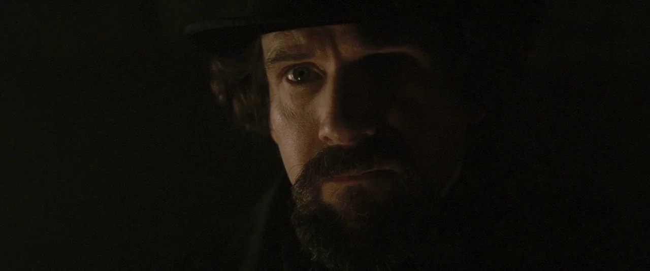 The Invisible Woman (2013) movie still: closeup — A dimly lit, dark space obscures the background environment. A man in a brimmed hat gazes forward…
