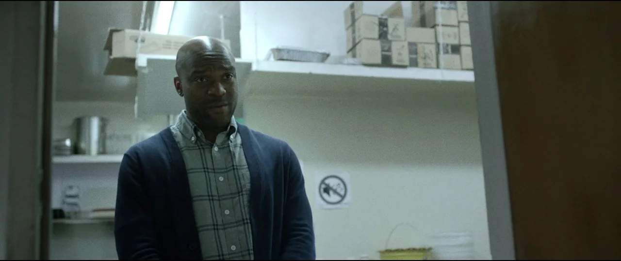 Lowlife (2017) movie still: medium — An interior storage room contains stacked cardboard boxes and a no-smoking sign on walls. A man in a blue…