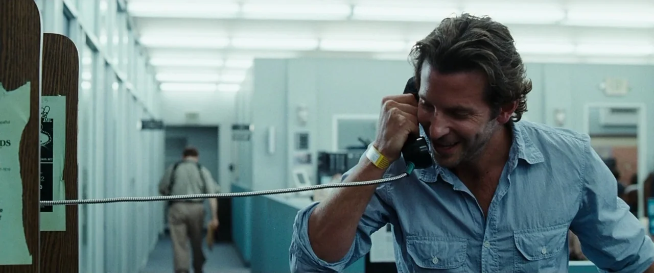 The Hangover (2009) movie still: medium — A man speaks on a telephone in the foreground of a sterile police office. In the background, a person in…