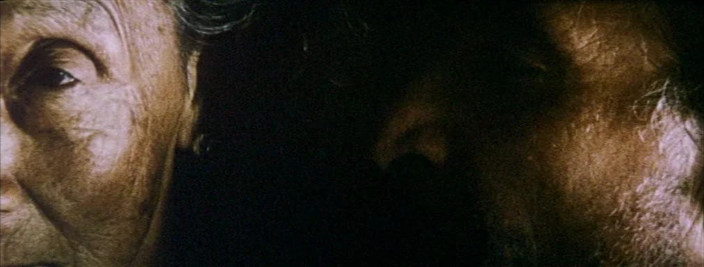 Japón (2002) movie still: closeup — Two faces are positioned on opposite sides of a dark and undefined interior space. The side lighting accentuates…