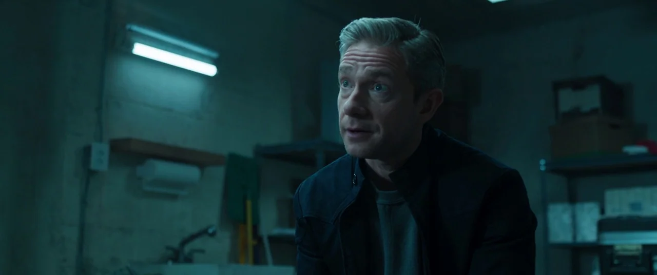 Black Panther (2018) movie still: medium — A dimly lit utility room features concrete walls and industrial metal storage shelving. A man in a dark…