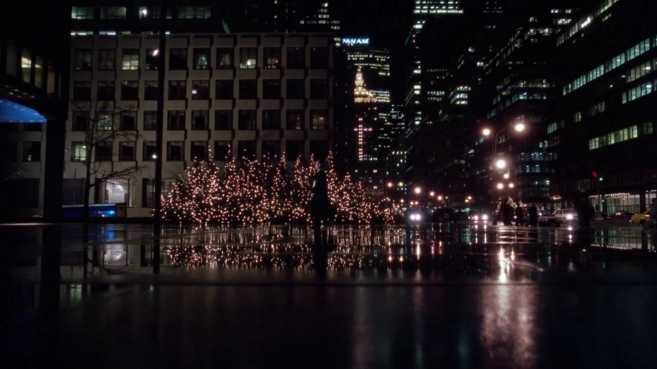 Scrooged (1988) movie still: wide — A dark, wet city plaza at night is illuminated by illuminated decorative evergreen trees. Reflections of the…