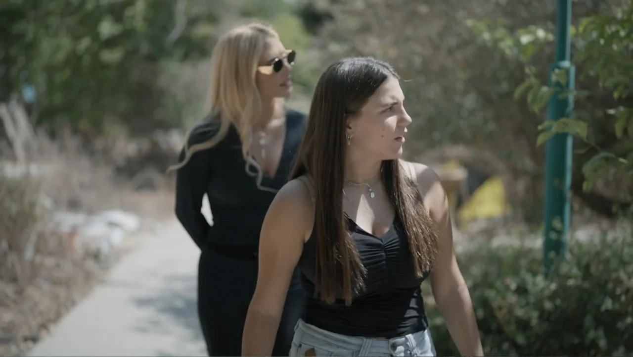 The Children of October 7 (2025) movie still: medium — Two women walk together along a paved walkway surrounded by greenery in a park. They move…