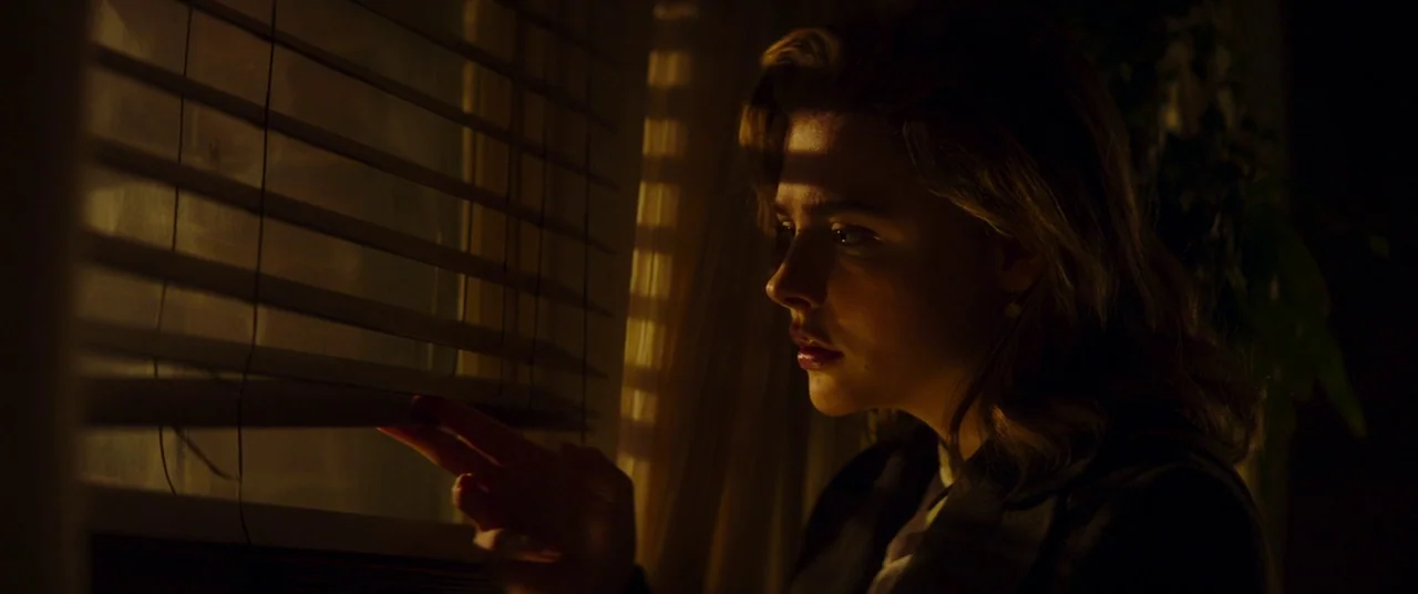 Greta movie still: closeup — A dark indoor space is lit by golden light filtering through horizontal window blinds. A young woman with blonde hair…