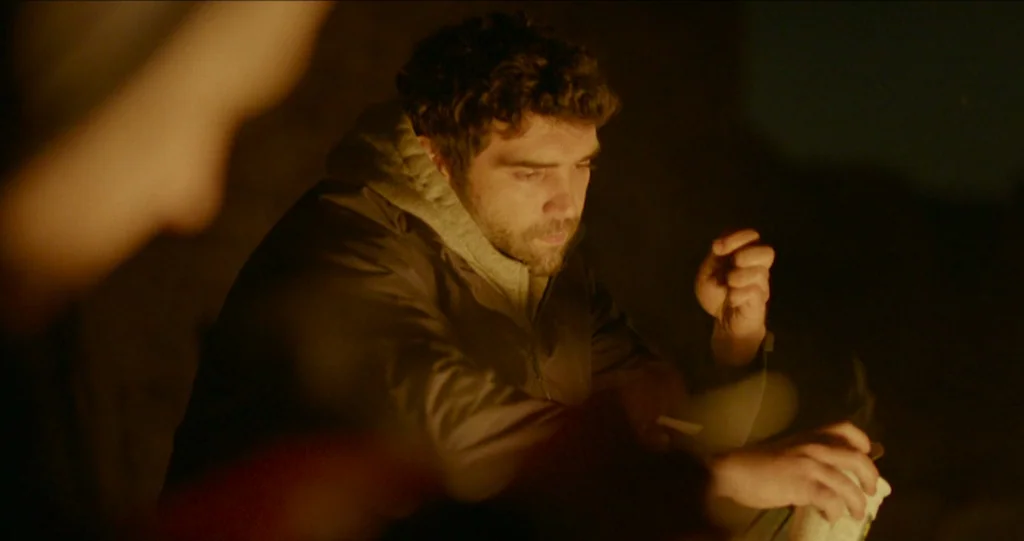 God’s Own Country (2017) movie still: medium — An outdoor night setting featuring a man sitting near a campfire. The subject holds a small object…