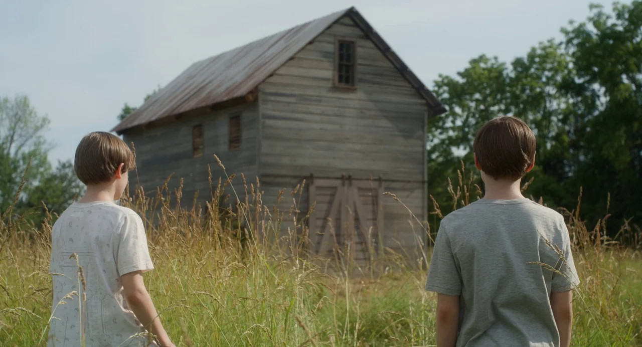 Goodnight Mommy (2022) movie still: medium — A field of tall dry grass surrounds two young boys facing a large barn. The weathered gray barn sits in…