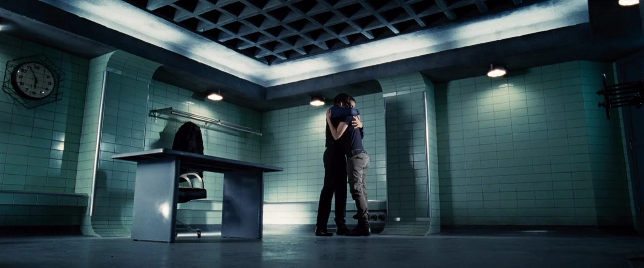 The Hunger Games (2012) movie still: wide — A wide, tiled detention room features a geometric ceiling grid and a stark metal desk. Two individuals…