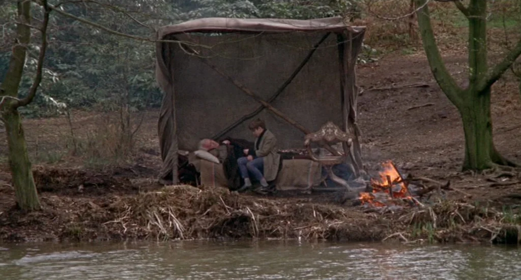 Fahrenheit 451 (1966) movie still: wide — A clearing by a riverbank features a crude canvas shelter constructed from wooden poles. A boy sits on a…