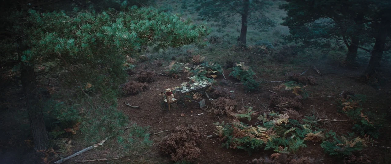Christopher Robin (2018) movie still: establishing — A clearing in a dense woodland features a wooden table and a small chair. A stuffed bear sits…