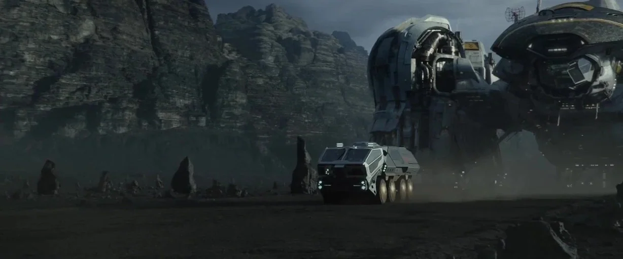 Prometheus (2012) movie still: wide — A rugged, dark planet landscape features sharp rocky formations under a cloudy grey sky. A small,…
