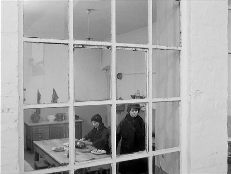 Effi Briest (1974) movie still: medium — A plain interior room with a wooden table and a large window frame. Two women are present, one working at…