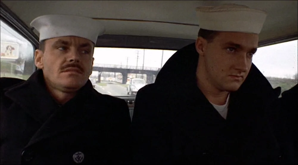 The Last Detail (1973) movie still: medium — Two men in navy uniforms sit in the back seat of a moving vehicle. Outside the window, a concrete…
